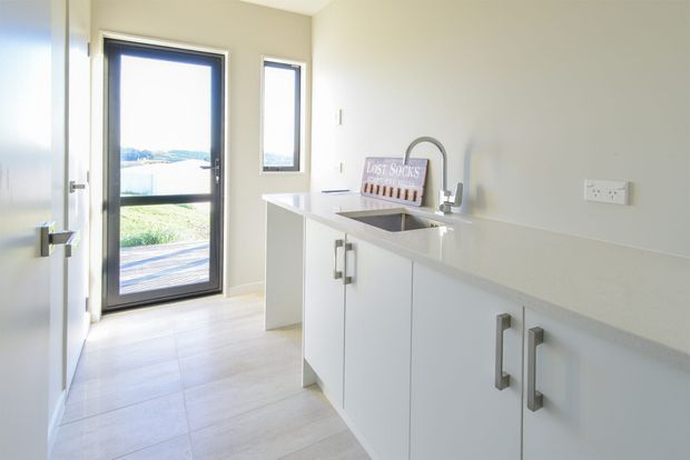 Bright, compact laundry room with white built-in cabinetry, a long white benchtop, stainless sink and gooseneck tap, pale tiled flooring, and dark-framed glazed exterior doors and windows. A small sign rests behind the sink, while the glazing provides views to the landscape outside.