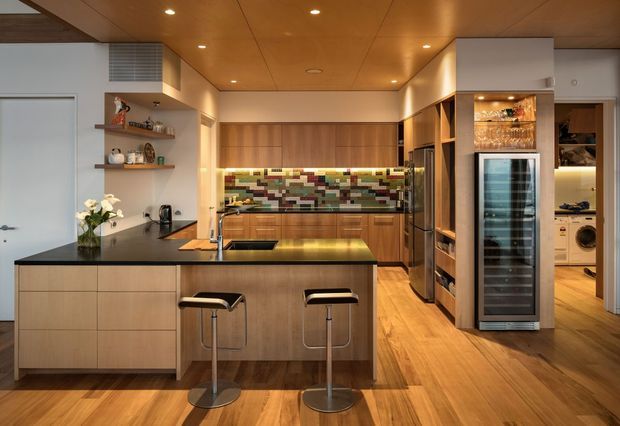 A warm, contemporary kitchen viewed straight on, featuring a timber island with dark stone benchtop, integrated sink and two stools. Full-height timber cabinetry lines the rear wall, with a colourful geometric mosaic splashback, open display shelving, built-in appliances, and a glass-front wine refrigerator. Timber flooring and a timber panelled ceiling create a cohesive, softly lit interior. A laundry area is visible through the doorway on the right.