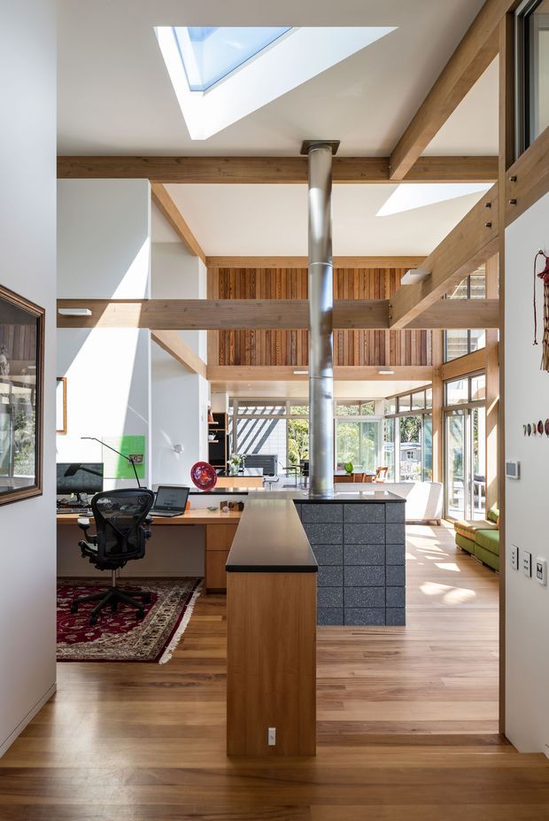 Bright open-plan interior viewed from a passage or entry, featuring a timber kitchen island with a dark benchtop, integrated cabinetry, a central stainless-steel flue, exposed timber beams, vertical timber wall panelling, skylights, and extensive glazing. A compact home workspace with desk and office chair sits to the left, while a green sofa is visible in the adjoining living area. Natural light creates strong geometric shadows across the timber floors.