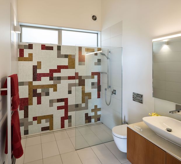 Bright contemporary bathroom with a walk-in glass shower, wall-hung toilet, floating timber vanity, vessel basin, and large illuminated mirror. The shower wall features a distinctive geometric mosaic in white, grey, red, ochre, and brown, while the remaining walls and floor are finished in light neutral tiles.