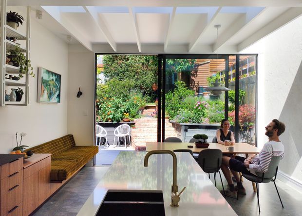 Bright open-plan kitchen and dining area with a white island, integrated sink and brass-toned tap, timber cabinetry, and a small dining table. Full-height black-framed glazing opens to a lush planted courtyard and brick steps, while roof glazing and white exposed ceiling beams bring in abundant natural light. A built-in timber bench, open shelving, artwork, and indoor plants add warmth.