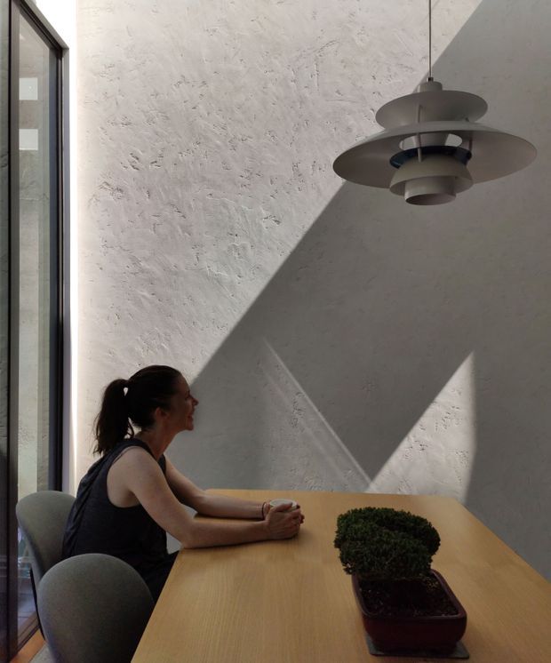 A contemporary dining area with a light timber dining table, rounded upholstered chairs, a sculptural grey pendant light, and a textured white plaster wall. Strong sunlight through the tall black-framed window creates dramatic geometric shadows across the wall. A small bonsai-like plant sits on the table, with a person seated at the table.