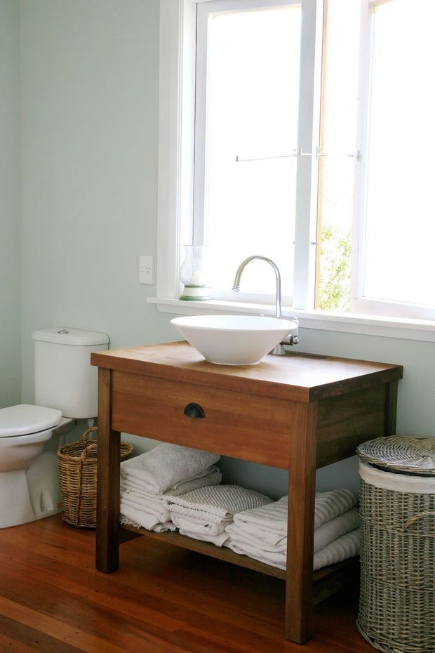 A bright, compact bathroom with pale blue-green walls, a white toilet, and a freestanding timber vanity featuring an open lower shelf, white vessel basin, and brushed metal faucet. Large white-framed windows provide abundant natural light. Woven baskets and neatly folded white towels add practical storage and a warm, natural touch.