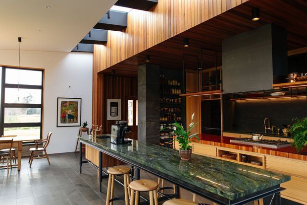 A spacious open-plan kitchen and dining area with a long dark green stone island, timber cabinetry, extensive vertical timber wall and ceiling panelling, dark structural columns, black-framed windows, skylights, and warm integrated lighting. A dining table sits near the windows, while open shelving, appliances, and a cooktop are integrated into the kitchen joinery.