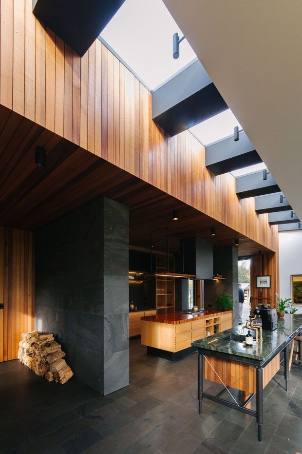A dramatic contemporary kitchen and living space with a tall linear skylight, exposed dark structural beams, and extensive vertical timber wall cladding. The kitchen features warm timber cabinetry, a dark natural-stone island benchtop, dark tiled flooring, black cabinetry and appliances, and a large dark stone fireplace volume with stacked firewood. Pendant and recessed lighting highlight the double-height interior.