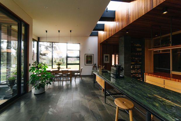 Open-plan contemporary interior combining a dining area and a richly timber-lined kitchen. Full-height glazing brings in abundant daylight, while dark stone-look flooring, a long dark green stone island with timber stools, black-framed windows, and warm vertical timber wall and ceiling cladding create a refined, natural atmosphere. The kitchen includes integrated display shelving and a dramatic double-height or partially voided timber feature zone.