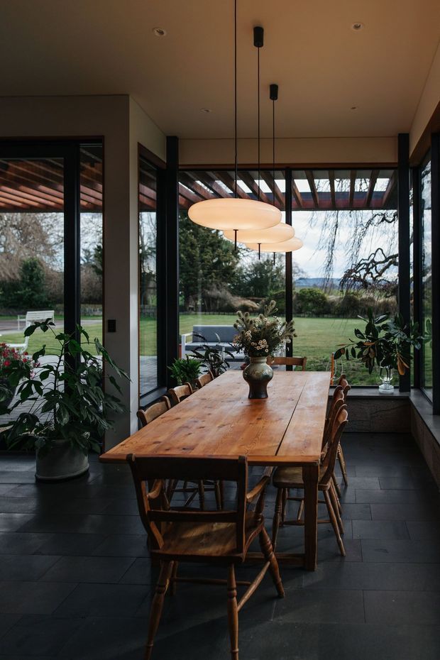 A warm, moody dining area with a long rustic timber dining table and matching wooden chairs, framed by extensive black-framed glazing overlooking a landscaped garden. Three sculptural oval pendant lights hang above the table, with potted plants and a central floral arrangement adding an organic feel. Dark tiled flooring and exposed timber pergola elements are visible through the glazing.