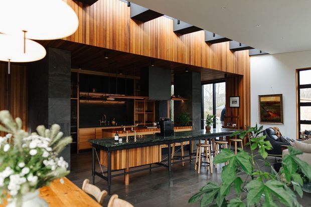 Open-plan contemporary kitchen and living area with a large island bench, dark stone surfaces, extensive vertical timber wall panelling and cabinetry, black structural accents, pendant lighting, and tall windows. The space includes bar stools, indoor plants, artwork, and a dining table in the foreground.