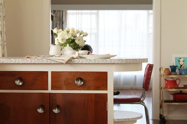 A warm, characterful dining and kitchen joinery scene featuring a dark timber built-in cabinet with round metallic pulls, a pale patterned stone or tile benchtop, and a compact dining table behind it. A large window with sheer curtains provides soft natural light, while a red dining chair and open shelving with colourful kitchenware add accents. The space has a refined mid-century-inspired feel.