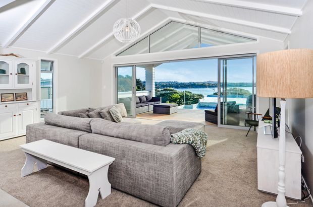 Bright, airy living room with a large grey sectional sofa, white coffee table, carpeted floor, white built-in cabinetry, and a vaulted white-panelled ceiling with exposed beams. Large sliding glass doors open to a balcony or terrace overlooking a coastal waterfront landscape, while triangular clerestory glazing and a spherical pendant enhance the room's openness.