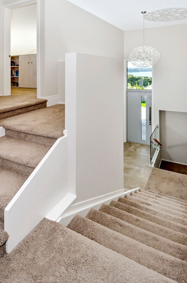 Bright contemporary stairway and landing with broad carpeted stairs descending toward a glazed entry door and a second carpeted flight leading upward. White plaster walls, white balustrades, and pale neutral carpeting create a clean, airy interior. A sculptural pendant light hangs over the stairwell, with a glimpse of built-in cabinetry in the upper hallway and outdoor greenery visible through the entry glazing.