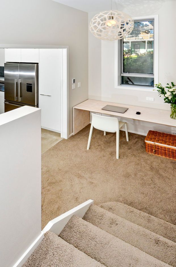 Bright contemporary open-plan interior viewed from a carpeted staircase, showing a compact kitchen with white cabinetry and stainless-steel refrigerator, a built-in timber-toned study desk beneath a window, white desk chair, woven storage ottoman, and a sculptural pendant light. The space uses soft neutral finishes and receives abundant natural light.