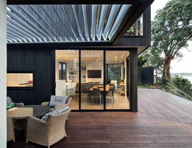 Contemporary dark-clad home opening onto a broad timber deck, with an outdoor dining setting in the foreground and full-height glazed doors revealing a bright dining and living area. A dramatic louvred roof canopy projects over the deck, while mature trees and a glimpse of water are visible beyond the property.
