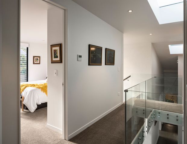 A bright contemporary upper-level hallway and landing with white walls, charcoal carpet, recessed downlights, and a large skylight. A frameless glass balustrade overlooks the lower level, while an open doorway reveals a bedroom with a white bed and mustard throw. Framed artwork and a wall control panel line the corridor.