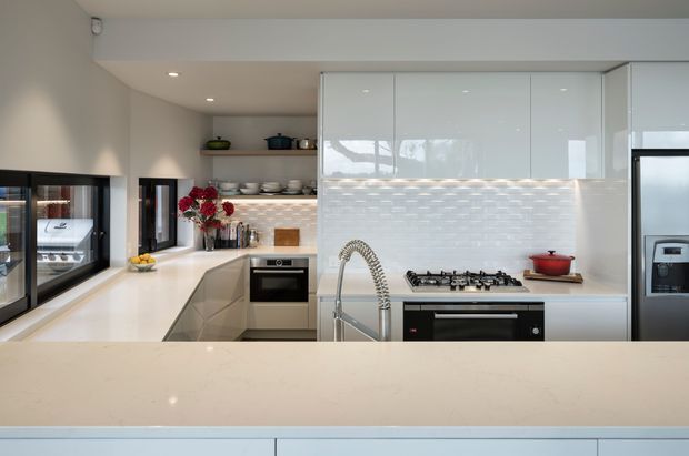 Bright contemporary galley-style kitchen viewed across a large pale stone island. The space features glossy white full-height cabinetry, white benchtops, a textured light mosaic-tile splashback, integrated stainless appliances, a gas cooktop, and a prominent spring-coil kitchen tap. A serving window connects to another room, while open shelving and a small floral arrangement add warmth.