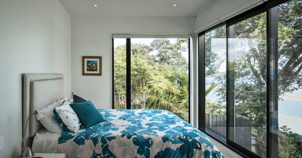 Bright contemporary bedroom with a pale upholstered bedhead, blue patterned bedding, and full-height black-framed glazing wrapping around two walls. The windows overlook lush vegetation and a body of water, creating an airy connection to the outdoors.