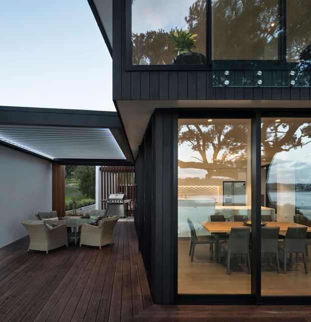 Modern two-storey house exterior with dark vertical cladding, large black-framed glazed doors and windows, and a timber deck. The covered outdoor entertaining area contains wicker lounge seating and a barbecue, while the glazing reveals a warm dining area inside. Water and trees are visible in reflections and beyond the property.
