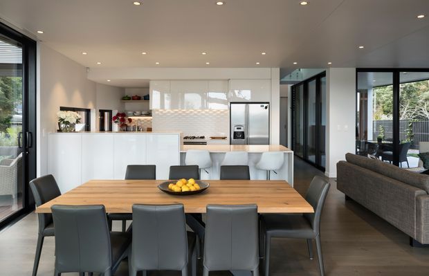 Open-plan contemporary kitchen, dining and living area with a large timber dining table, dark upholstered dining chairs, a white island bench, glossy white cabinetry, stainless-steel appliances, and a tiled geometric splashback. Floor-to-ceiling glazing and dark-framed doors provide abundant natural light, while a grey sofa sits in the adjoining living zone.