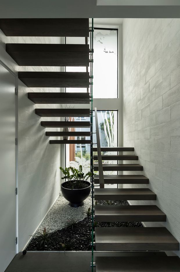 A dramatic modern interior stairway with floating dark timber treads, a slender central support, and clear glass balustrades. The stair rises through a narrow, tall void between textured pale walls, with large vertical windows bringing in daylight and a gravel-filled internal garden with potted greenery below.