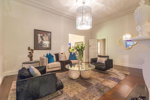 Elegant formal living room with cream walls, decorative ceiling mouldings, dark timber flooring, a patterned area rug, and a refined seating arrangement of sofas and armchairs around a round glass coffee table. The room features a crystal pendant light, framed artwork, sculptural decor, and a decorative mirrored wall piece with blue and gold accents.