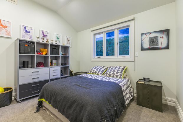 A compact, bright bedroom with a double bed, patterned geometric pillows and bedspread, dark grey throw, carpeted floor, built-in-style shelving and drawer storage, framed artwork, and a white-framed window overlooking greenery. The room has a clean contemporary appearance with a restrained grey, white, and green palette.