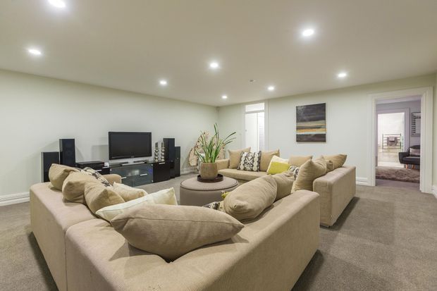 A spacious, softly lit living room with a large beige U-shaped sectional sofa arranged around a round coffee table. The room has neutral painted walls, grey-beige carpet, recessed ceiling lights, a wall-mounted television and media console, indoor plants, and framed artwork. An open doorway reveals an adjacent bedroom.