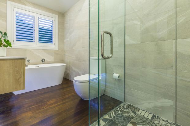 Bright contemporary bathroom with a freestanding white bathtub, wall-mounted timber vanity, white toilet, frameless glass shower enclosure, patterned shower floor tiles, large pale stone-look wall tiles, and dark timber flooring. A shuttered window provides natural light, with a small plant adding greenery.