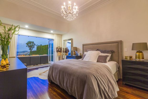 Elegant, spacious bedroom with a large upholstered bed, layered neutral bedding, dark bedside cabinetry, polished timber flooring, ornate ceiling moulding, and a crystal chandelier. Full-height glazing opens toward an outdoor terrace or balcony, bringing in evening light and views. The room has a refined, luxurious transitional character.