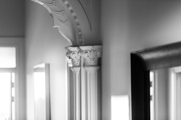 Close-up black-and-white interior view highlighting ornate classical plasterwork: a fluted pilaster with acanthus-leaf capital, decorative arch moulding, and a dark-framed mirror at right. Bright window openings are visible in the background.