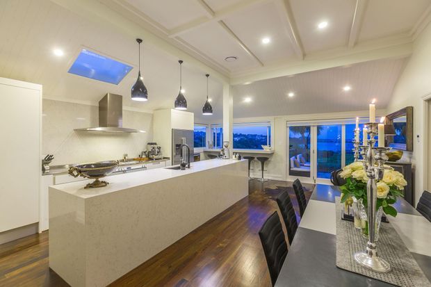 Bright open-plan kitchen and dining area with a large white stone island, integrated cabinetry, pendant lights, dark timber flooring, and a coffered ceiling. The adjoining dining space opens to glazed doors with a blue-hour exterior view.