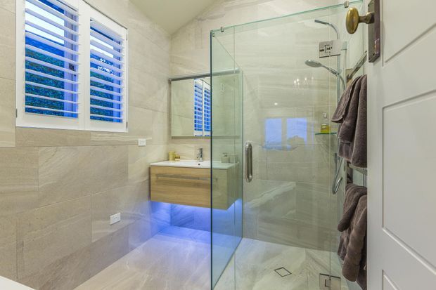 Bright contemporary bathroom featuring a frameless glass walk-in shower, large-format beige stone-look wall and floor tiles, a floating timber-look vanity with under-cabinet lighting, white plantation shutters, and brushed metal shower fittings. A partially visible white door and towel storage are on the right.