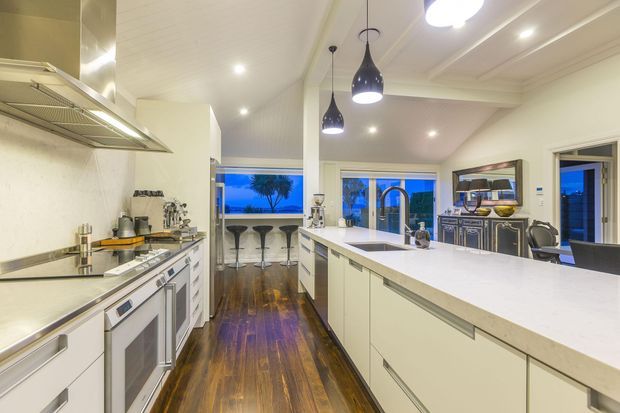 A bright, spacious contemporary kitchen viewed from the cooking zone toward a large central island and adjoining breakfast bar. The room features white shaker-style cabinetry, pale stone benchtops, stainless-steel appliances, a timber floor, pendant lights, and a coffered white ceiling. Large windows and a glazed door provide views and abundant evening light.
