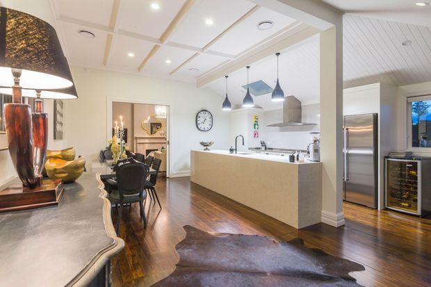 Open-plan kitchen and dining area with a large central island, stainless-steel appliances, pendant lighting, dark timber flooring, and a coffered ceiling with exposed decorative beams. The adjoining dining space features a dark dining table and chairs, while a sculptural console and table lamp occupy the foreground.
