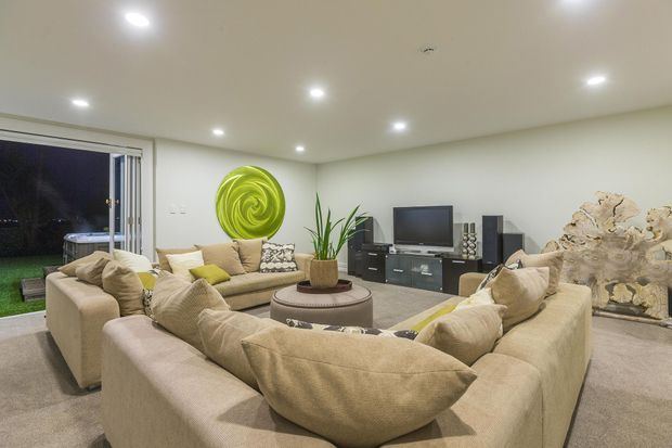 A spacious contemporary living room with two large cream upholstered sofas arranged around a round coffee table. The room has recessed ceiling lights, pale walls, neutral carpet, a wall-mounted green circular artwork, indoor plants, a television and low media cabinetry. Large sliding glass doors open toward a dark outdoor area, while a sculptural pale timber feature adds visual interest near the media wall.