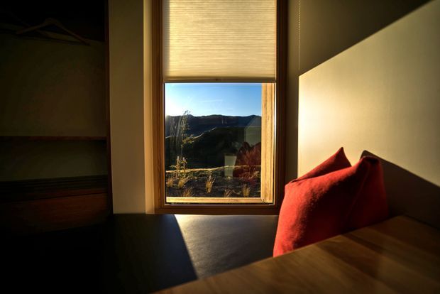 A warmly lit bedroom corner with a built-in dark upholstered window seat, vivid red cushions, and a timber-framed window overlooking a rugged mountainous landscape. A partially visible built-in wardrobe or shelving unit occupies the left side, while strong sunlight creates dramatic contrast across the pale wall and seat.