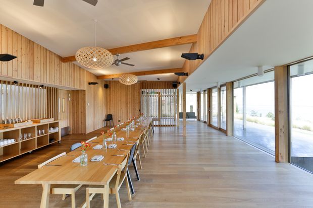 A spacious open-plan dining and communal gathering space with a long timber dining table set for a meal, timber wall panelling, exposed timber beams, woven pendant lights, and extensive floor-to-ceiling glazing along one side. The room has a warm natural material palette and views toward an outdoor landscape.