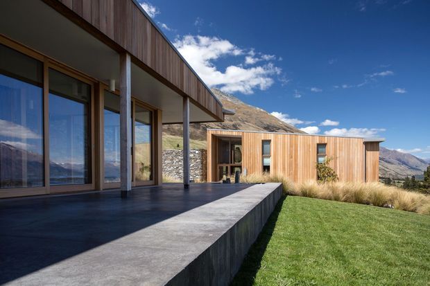 Contemporary single-storey mountain residence with a low-pitched roof, extensive floor-to-ceiling glazing, timber-clad walls, and a broad concrete terrace overlooking a rugged alpine landscape. The architecture combines strong horizontal lines, slender columns, and natural materials in a clean, minimalist composition.