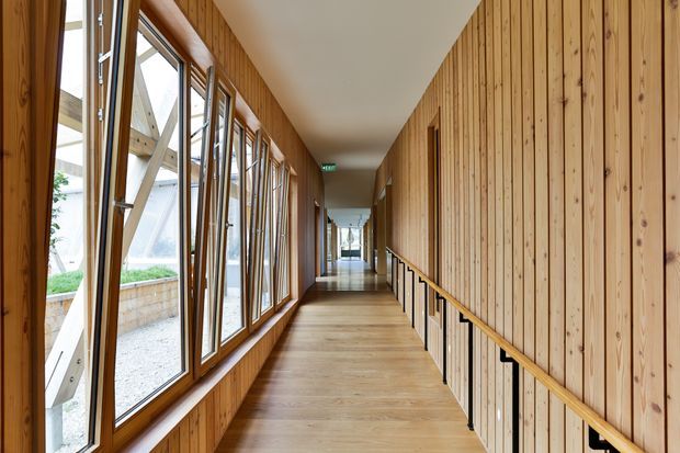 A long, narrow timber-lined corridor with warm vertical wood wall panelling, matching timber flooring and handrails, and a continuous row of tall windows along one side. The glazing overlooks a light-filled exterior courtyard or covered structure, while a simple black balustrade lines the opposite side. The space has a clean, contemporary architectural character with strong linear perspective.