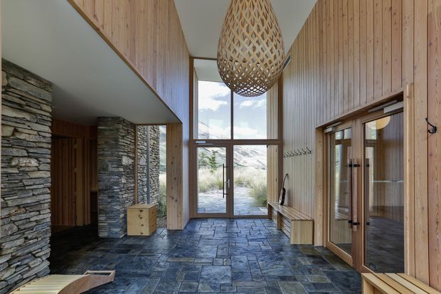 A tall, light-filled residential entry hall with double-height glazing and a central glass door opening to an outdoor landscape. The space features vertical timber wall panelling, extensive stacked natural-stone walls and columns, dark slate-like floor tiles, timber-framed glazed doors, built-in timber bench seating, and a large woven pendant light. A secondary glazed room is visible through the doors on the right.
