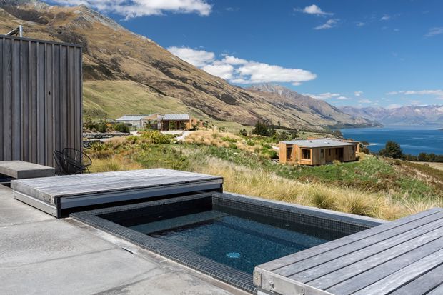 A contemporary hillside residential setting overlooking a blue alpine lake, with a compact dark-tiled swimming pool integrated into a concrete terrace. Timber-clad buildings sit among native grasses on the sloping site, backed by rugged mountains and expansive sky.