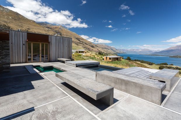 A contemporary rooftop or elevated outdoor terrace with concrete seating and paving, a small rectangular plunge pool, timber-clad pavilion with glazed doors, and a dramatic lakeside mountain setting. The space has a minimalist, geometric design with expansive views across the blue lake.