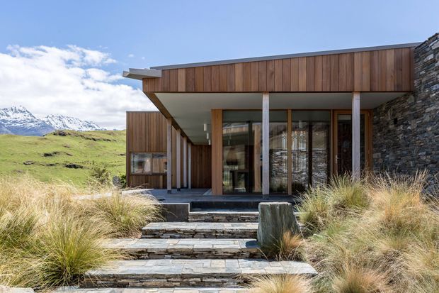 A contemporary single-storey mountain home with a flat roof, extensive timber cladding, floor-to-ceiling glazing, exposed concrete columns, and a rugged stacked-stone wall. Broad stone steps lead through native grasses to the recessed entrance, with snow-capped mountains visible beyond.