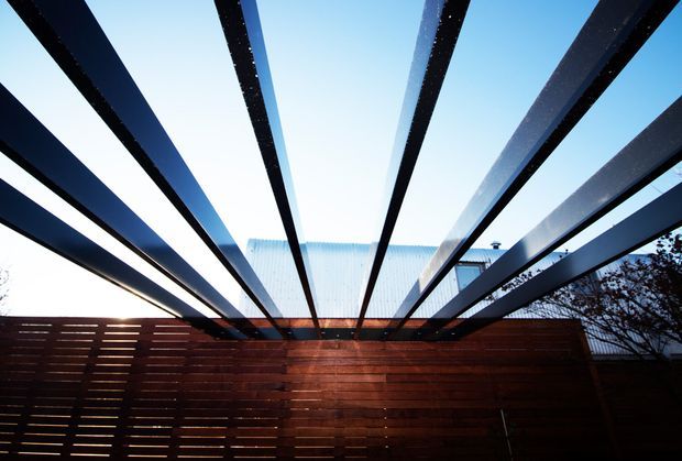 Exterior architectural detail viewed from a timber-fenced courtyard or outdoor area, featuring dramatic dark steel beams projecting overhead against a bright sky and a corrugated metal roof in the background. The horizontal timber screening creates a warm contrast with the black structural framework.