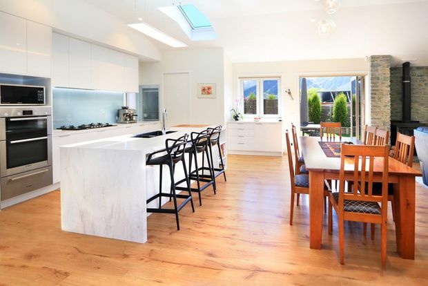 Bright open-plan kitchen and dining area with white flat-panel cabinetry, a large white stone island with black bar stools, integrated appliances, timber flooring, and a timber dining table beside garden-facing windows and doors. A skylight and generous glazing provide abundant natural light, while a stone-clad structural column and dark fireplace add contrast.