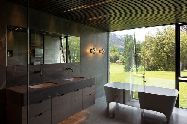 A sophisticated contemporary bathroom with a double vanity, wall-mounted taps, large mirror, freestanding bathtub, dark stone-look wall surfaces, and wood-toned built-in cabinetry. Full-height glazing opens the room to a manicured garden and dramatic mountain scenery. A dark linear slatted ceiling and circular wall sconces add architectural and decorative detail.