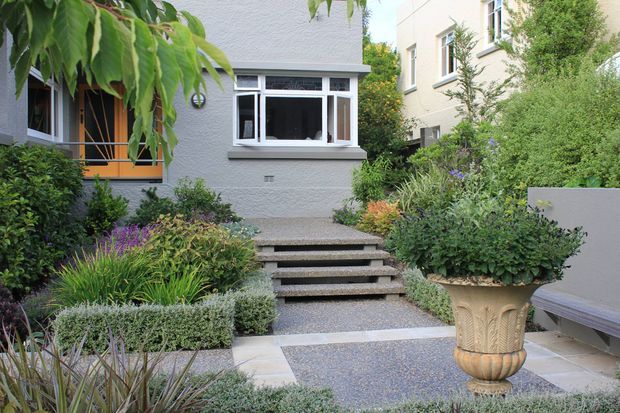 A landscaped residential courtyard or front garden leading to a raised house entrance. The scene features clipped shrubs, ornamental grasses, purple flowering plants, textured paving and gravel, concrete steps, a grey textured facade, white-framed windows, and a decorative urn planter. A bench is positioned at the right edge.