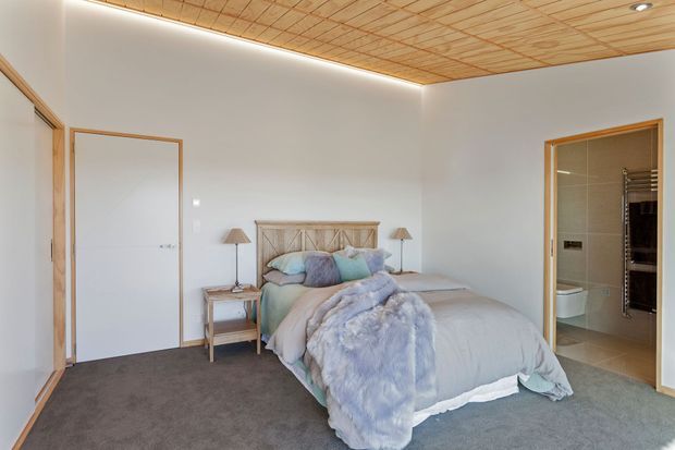 A bright, softly furnished bedroom with a large upholstered bed, pale blue and grey bedding, timber bedside tables, and a timber-framed headboard. The room has grey carpet, white walls, a warm timber grid-panel ceiling with concealed perimeter lighting, and timber-framed doors opening to a tiled bathroom.