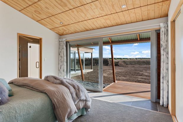 A bright bedroom with a low-pitched timber-lined ceiling, large sliding glass doors opening onto a timber deck, and views across a landscaped outdoor area. The room includes a partially visible upholstered bed with layered neutral bedding, patterned curtains, carpeted flooring, and a timber-framed doorway to an adjoining space.