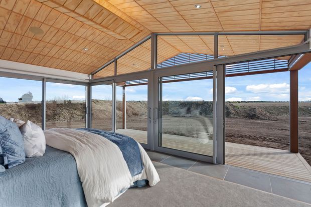 A bright contemporary bedroom with floor-to-ceiling glazing opening onto a timber deck and expansive rural landscape. The room features a pitched, timber-lined ceiling with exposed framing, grey metal-framed sliding doors and windows, large-format tiled flooring at the glazed edge, and a soft upholstered bed with blue and cream bedding.