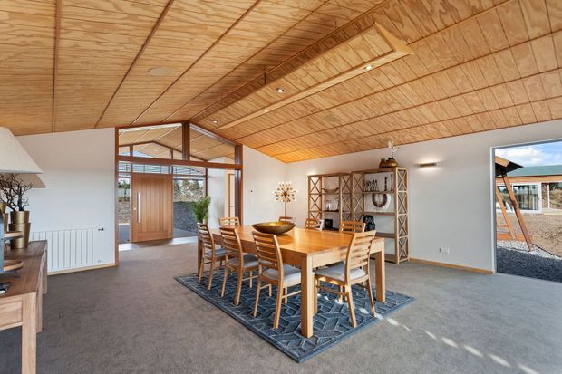 Open-plan dining area with a large timber dining table and matching chairs, set on a patterned rug. The room features extensive exposed timber plywood ceiling and wall cladding, white walls, grey carpet, timber-framed glazing and doors, and open shelving. A glazed entry and views to the exterior are visible beyond.